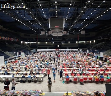 An indoor sports arena filled with rows of tables, each occupied by people engaged in competitive activities. The ceiling is high, with numerous lights illuminating the large space. The audience seating on either side is empty, and there are flags hanging from the upper level.