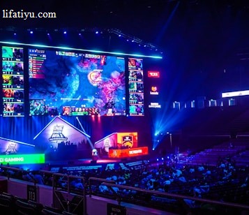 A large esports arena featuring a massive screen displaying a video game, surrounded by sponsor logos and team names. The auditorium is filled with spectators watching the gaming event. Neon blue and purple lights illuminate the stage, creating a vibrant atmosphere.