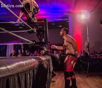 A wrestling match taking place in an indoor venue, where one participant is crouched on the top rope of the ring, while the other stands outside the ring looking at them. A crowd watches the event enthusiastically from their seats.