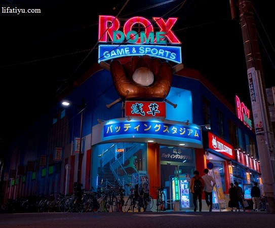 A vibrant, neon-lit facade of a gaming and sports establishment at night, featuring a large glove holding a baseball above the entrance. The building is adorned with colorful signs in both English and Japanese, with various advertisements and an illuminated sign that says 'ROX DOME'. Several people are walking towards the entrance, and bicycles are parked along the side.