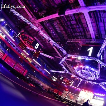 An indoor arena with vibrant purple and blue lighting, featuring large numbered screens suspended from the ceiling. The venue has a high ceiling with intricate metal truss structures and hanging lights, creating a futuristic and energetic atmosphere.