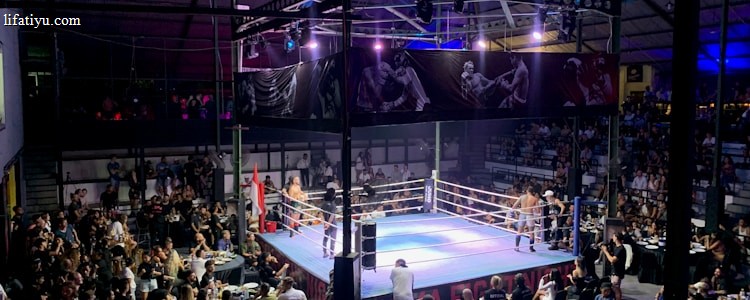 A crowded indoor arena with a boxing ring at the center, surrounded by an audience seated at tables. The scene is illuminated by overhead lights, casting a vibrant glow over the ring where two fighters are preparing for a match. Spectators are engaged, capturing the event with cameras and enjoying the lively atmosphere.