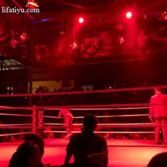 A wrestling ring is illuminated with red lighting, creating a dramatic atmosphere. Several people are visible around the ring, with silhouetted spectators in the foreground. Banners or art adorn the upper walls, and the venue has an industrial ceiling with exposed beams.