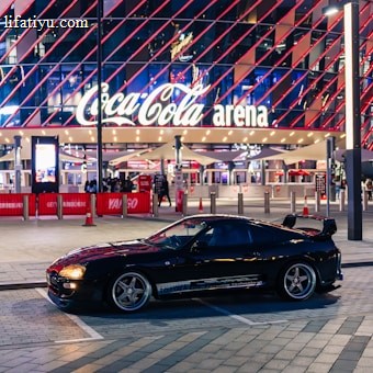 A sleek black sports car is parked in front of an illuminated arena entrance, with a large sign bearing the name of a famous beverage brand. The building features a modern, geometric facade with bright red and blue lights, creating a vibrant and lively atmosphere.